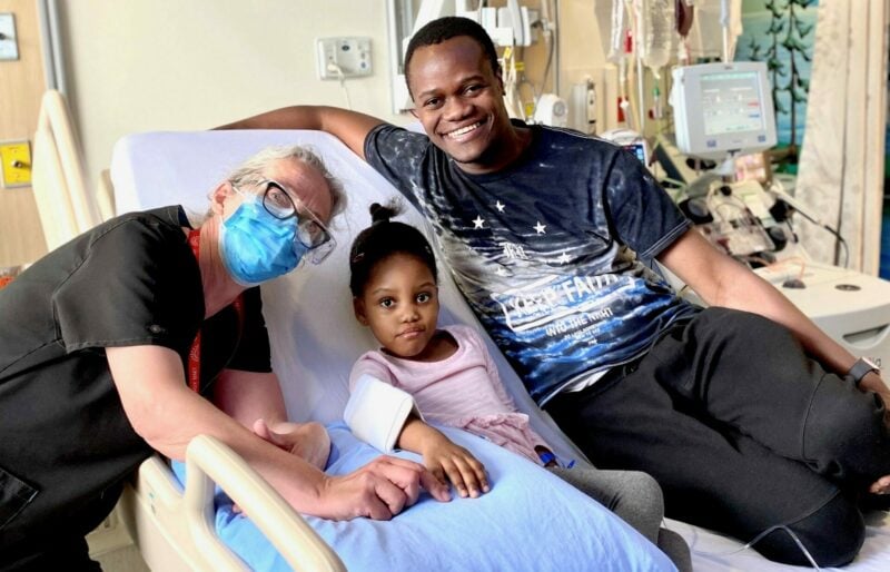 Young patient sits on hospital bed with father and attending nurse smiling on either side