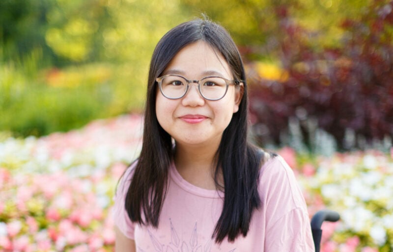 Amy wears a pink shirt and jeans in wheelchair in front of pink flower garden