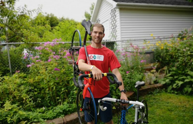 Young man stands smiling in a garden with a bike on his shoulder