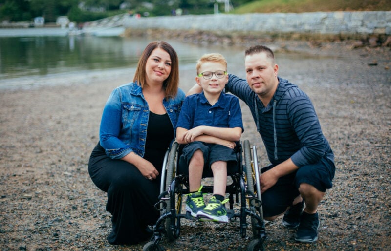 Owen is in wheelchair with his parents kneeling on both sides of him on a pebbled beach