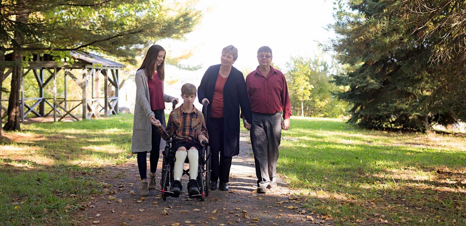 Patrick is in his wheelchair in a forest surrounded by his older parents and sister, holding his hand