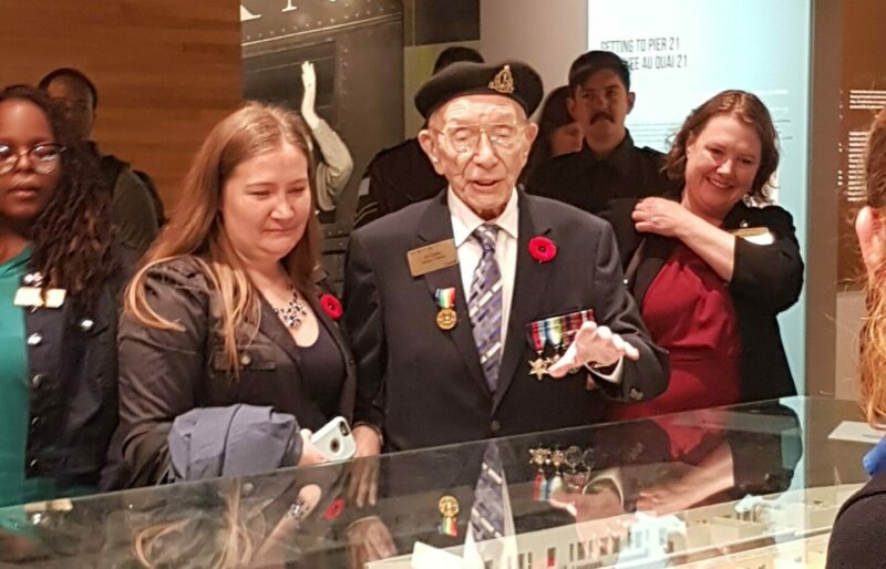 A veteran stands in front of a model of the Pier 21 immigration shed in its glass case with his granddaughter by his side.