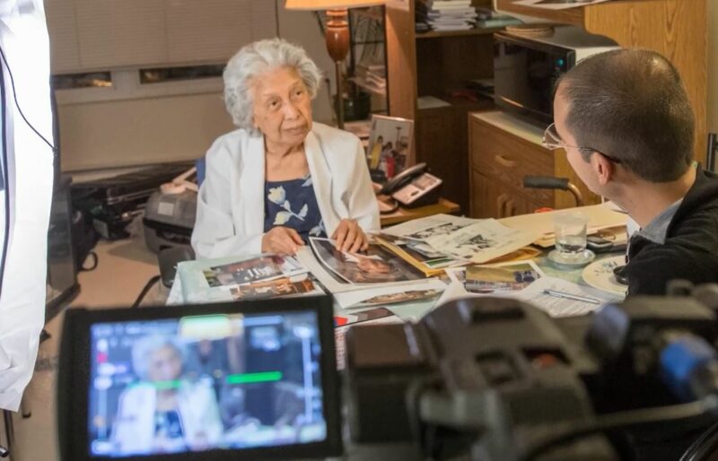 A woman stands at a desk with photos gathered in front of her as she speaks to an Oral Historian from the Museum who is facing her. The conversation is being recorded with a video camera.