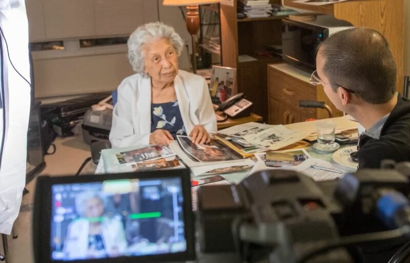 A woman stands at a desk with photos gathered in front of her as she speaks to an Oral Historian from the Museum who is facing her. The conversation is being recorded with a video camera.