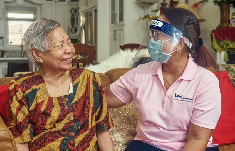 A woman wearing a face shield and a mask with her arm around an elderly Asian woman who is looking at her and smiling.