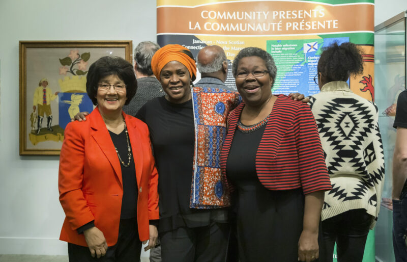 Three women stand closely together smiling at the camera in front of an exhibit. Three visitors are in the background reading the exhibit information.