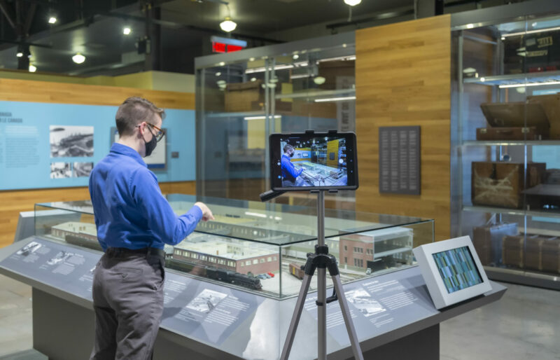 A Heritage Interpreter stands in front of the Pier 21 immigration shed model while an iPad captures this presentation.