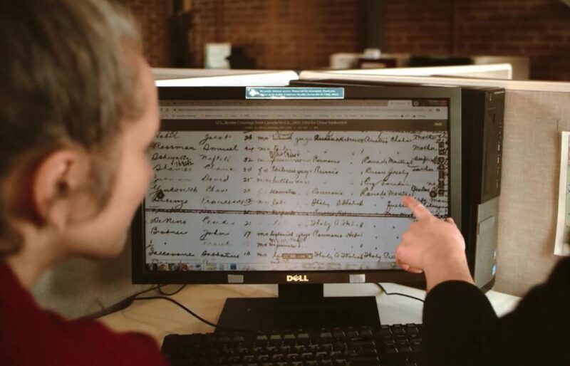 A woman faces a computer screen showing an immigration record of her family while one of our researchers points to a specific word on the screen during a genealogy family research.