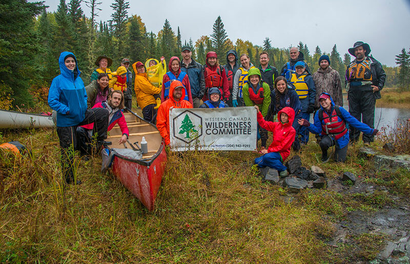 A group of people gather on a grassy slope in their raingear, canoe and Wilderness Committee sign in the fore.