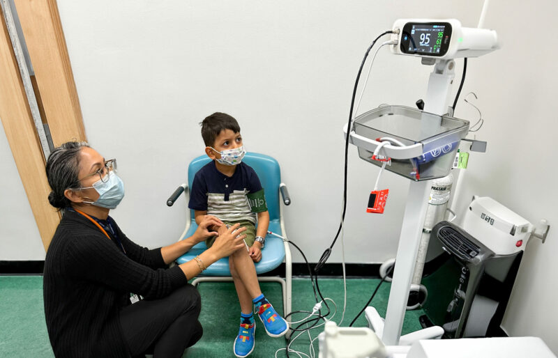 Simon is sitting on a chair while connected to a medical device, and a person with glasses and a mask over their mouth kneels next to him.