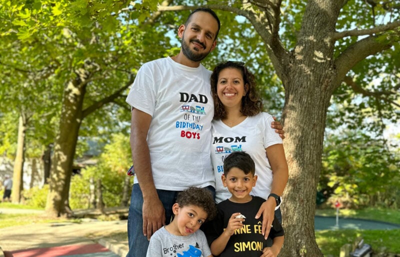 Simon is smiling outdoors in front of some trees. He is standing next to another smiling child beside him and both his father and mother directly behind him.