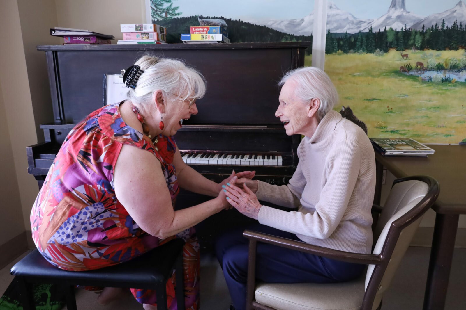 Two elderly women sitting at a piano laughing together while holding hands after playing a song.