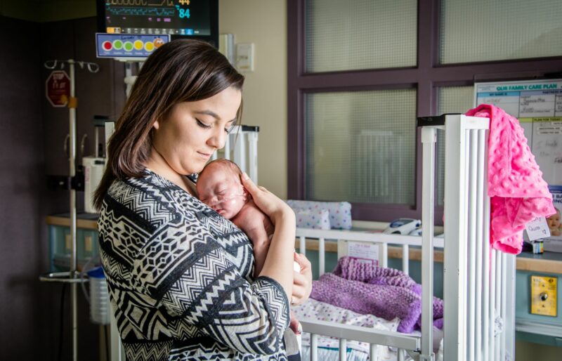 A mom holds her baby close to her chest in the NICU. She is wearing a black and white sweater and has brown hair. Behind them, you can see the baby's hospital bassinet in the background.