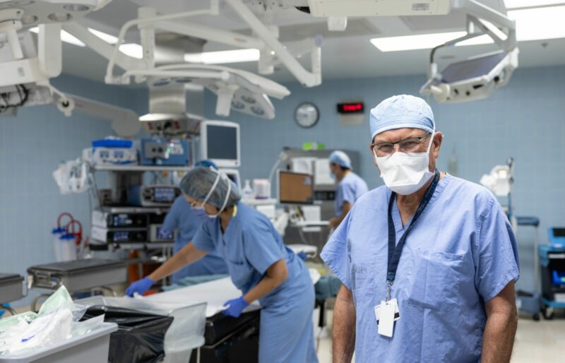 A health care work in a mask, scrubs and scrub cap is the focus of the image. In the background, you can see the operating room and other health care workers setting up