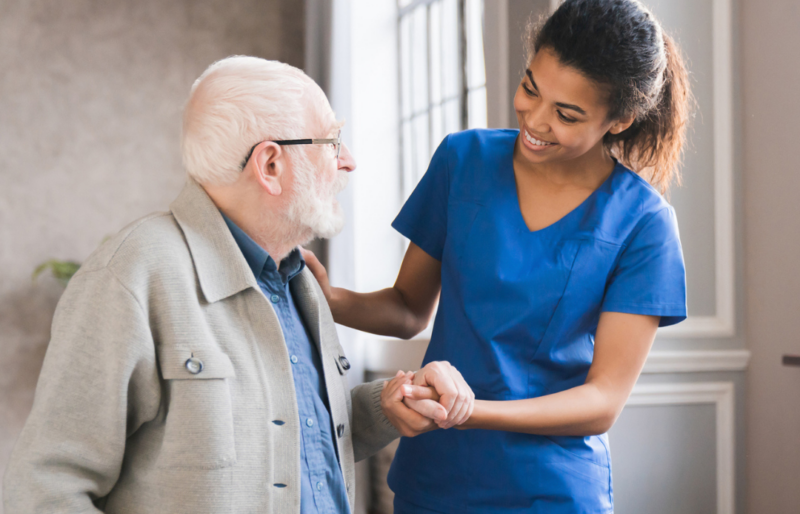 A female health care worker holds the arm of an elderly man. They look at each other as she offers him help. The man has white hair and white facial hair; he wears glasses a blue shirt and a tan jacket; you can see a cane in his right hand. The woman has brown hair pulled back in a ponytail and wears blue scrubs.