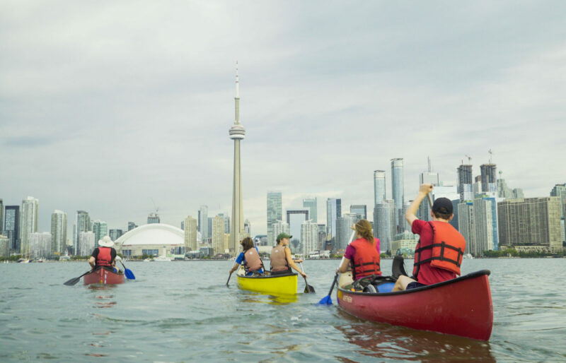 Image of three canoes paddling towards the Toronto skyline