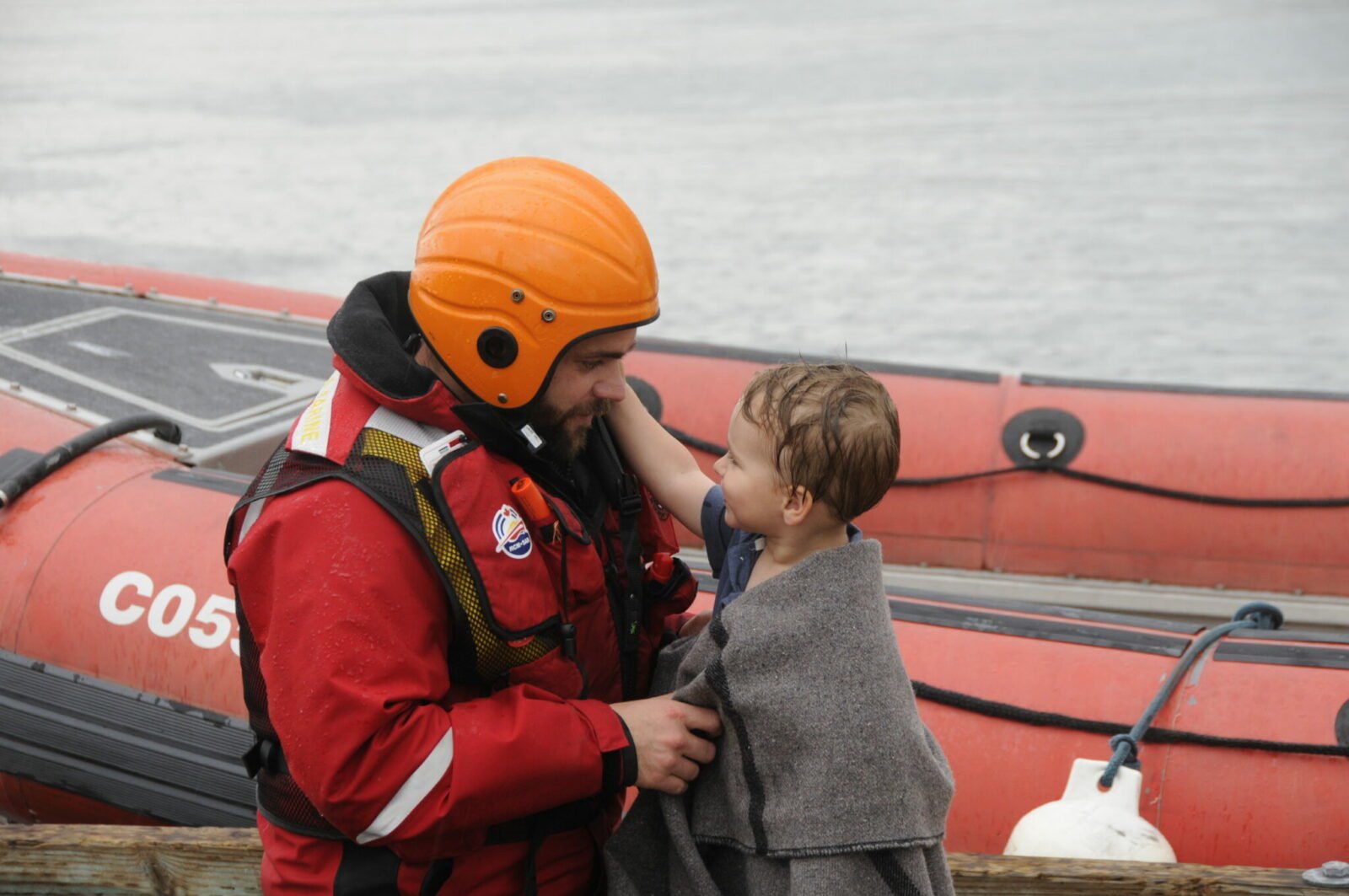 A marine search and rescue volunteer smiling places a blanket over a small wet child after rescuing him from the nearby ocean waters. The behind the two individuals is a red Royal Canadian Marine Search and Rescue vessel.