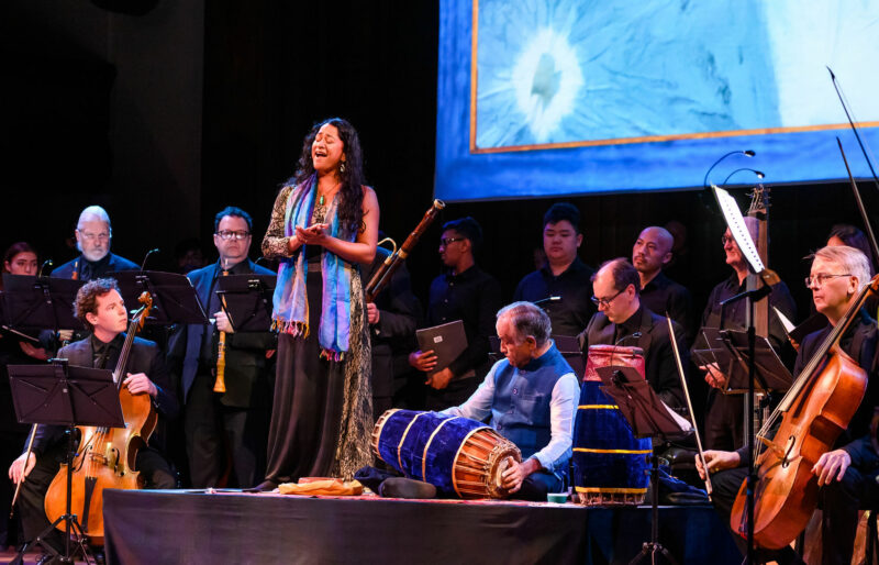 A singer stands on stage surrounded by standing and seated musicians