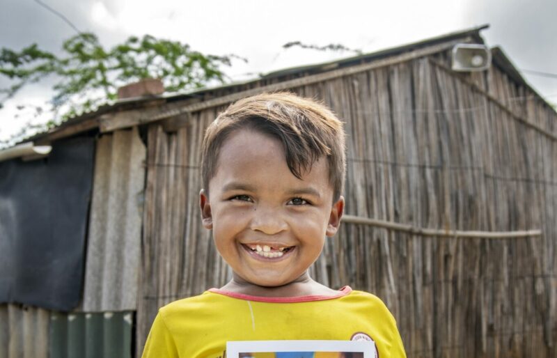 Boy in yellow shirt smiles as he holds a photo of himself before his cleft repair surgery.