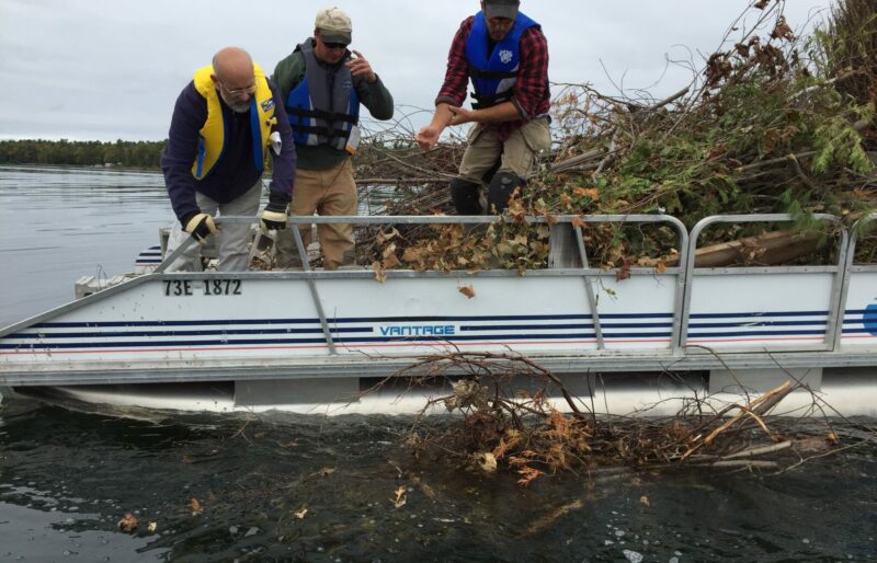 Three people on a boat dump brush bundles into a lake to restore underwater fish habitat.