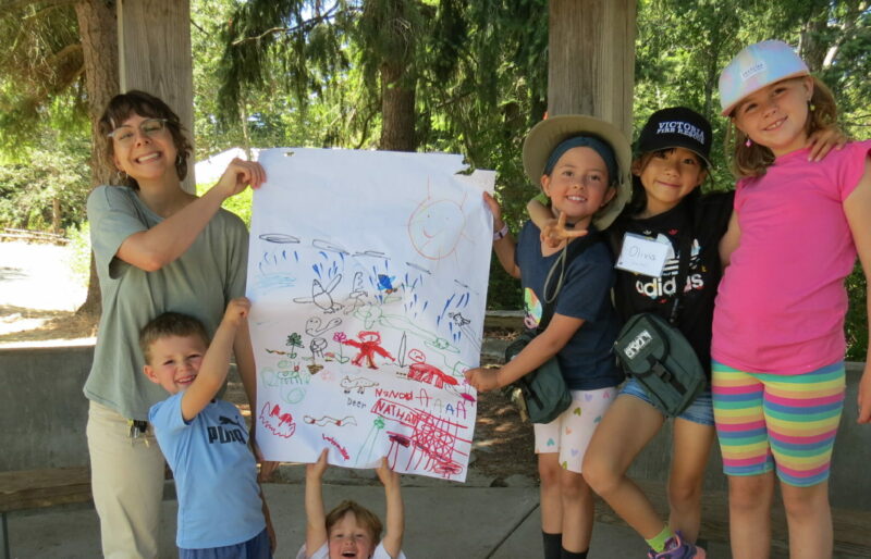 Five children, dressed in colourful summer attire, stand in a row. In the center there is a poster they have created. Two children on either side hold the poster up, and one kneels below it. On the far left, a woman with short, brown hair, a green shirt, and beige pants stands smiling.