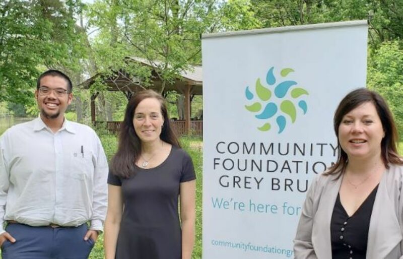 Three people smiling for a picture outside, in front of a Community Foundation Grey Bruce Banner