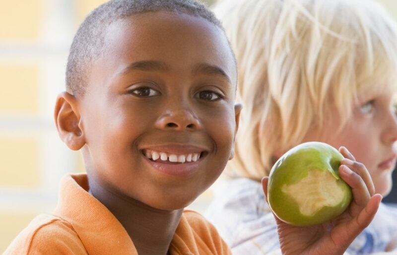 Young black boy holding a green apple with a couple of bites out of it. Looking at camera, smiling.