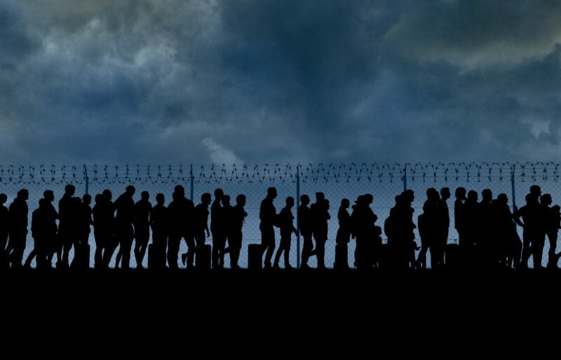 Line-up of migrants near the USA border against a fence with barbed wire.
