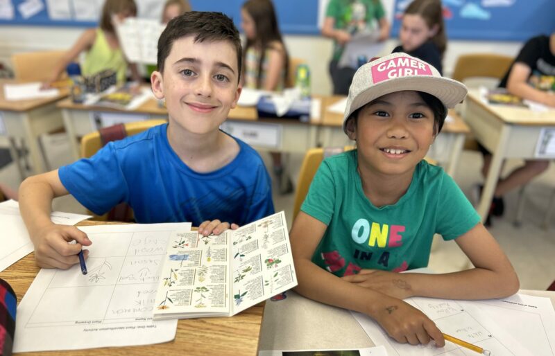 Two children sit at a desk working on an activity about native wildflower species in their area.