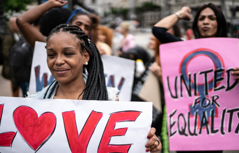 Transgender females holding signs during a demonstration in the street. Signs read 'Love' with a heart and 'United for equality'.