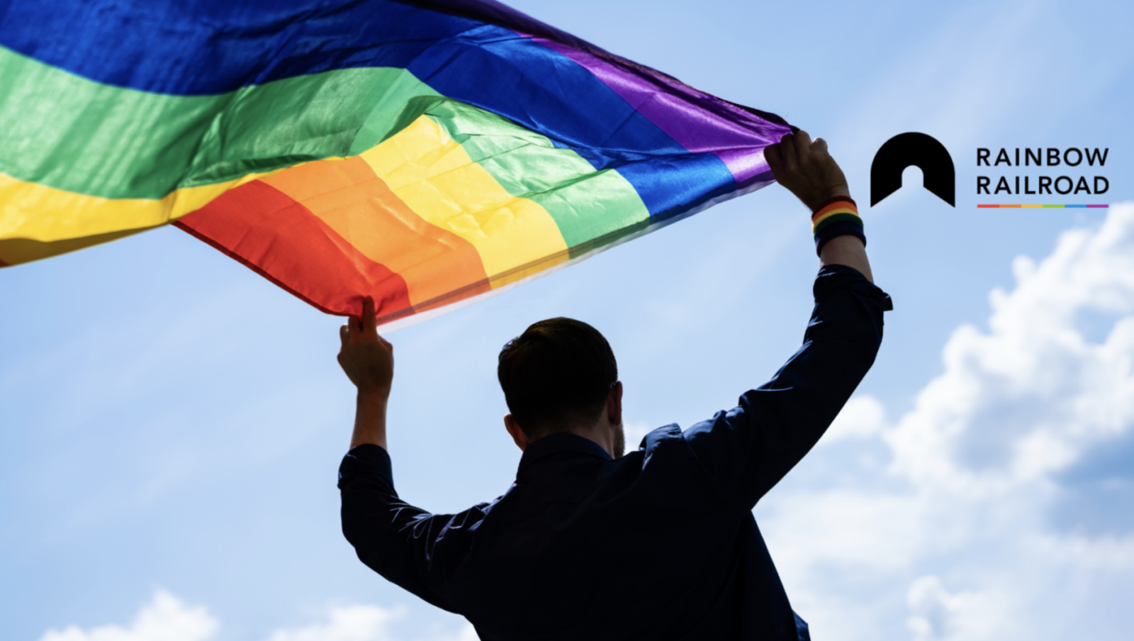 A person holds a rainbow flag over their head in the sky, with a Rainbow Railroad logo in the top-right corner.