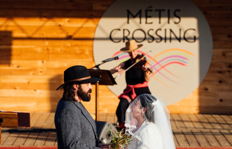 Two actors and a musician are standing on an outdoor stage on a sunny day. The actors are holding hands and facing each other. One actor is wearing a white wedding dress and the other actor has on a black cowboy hat, grey suit jacket and brown pants. A musician plays the fiddle in the background in front of a sign that reads Metis Crossing.