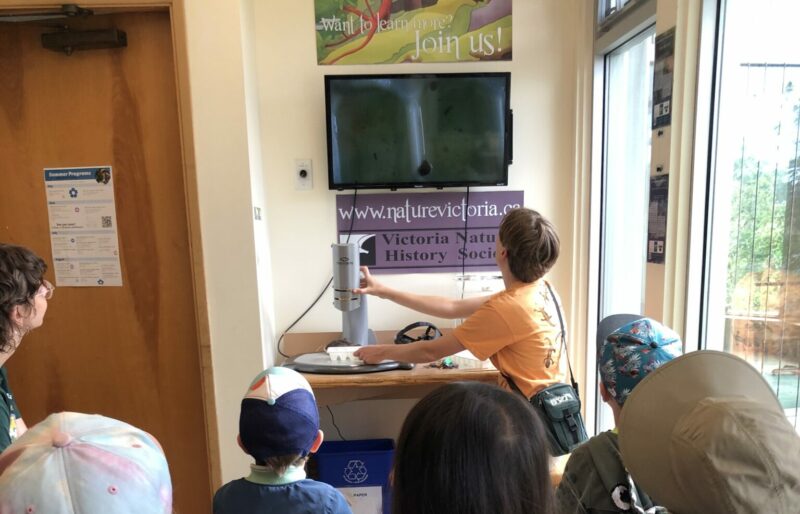 Children, dressed in summer attire, sit in two rows facing away from the camera. They are all looking up at a TV screen, which has a magnified water droplet on it. The instructor, with short hair and an orange shirt, looks up at the screen as well.