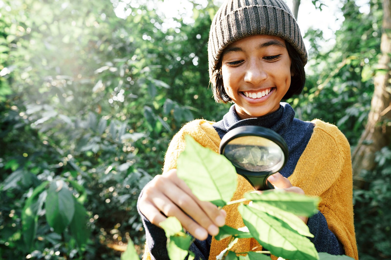 Learner in deciduous forest looking at a leaf with a magnifying glass. The learner has dark hair and a toque and a yellow sweater.