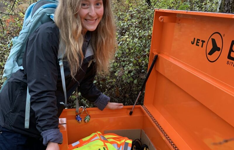 A blonde woman with wavy hair stands smiling over a big orange toolbox. The toolbox is propped open and in it you can see high visibility vests, large and small pruners, and a box of yellow and black work gloves.
