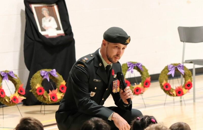 A uniformed officer holding a microphone speaks to a gymnasium of students.