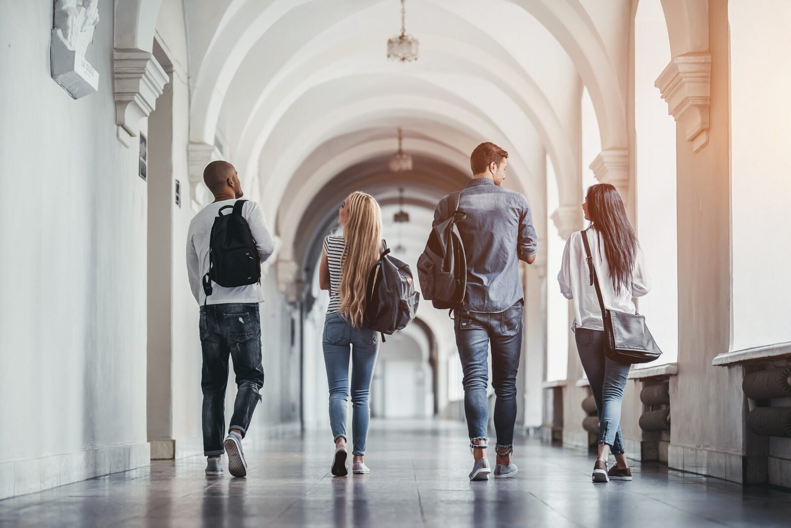 Four students with backpacks walking down a bright university hallway lined with arches.