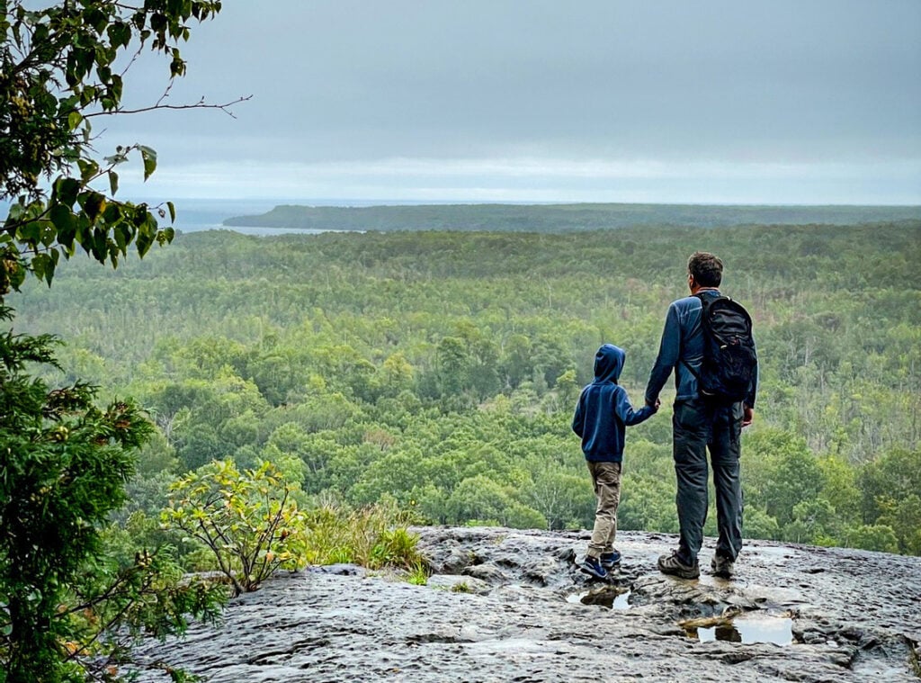 Father and son standing on a rock outcrop overlooking a forest and lake along the Bruce Trail, symbolizing protecting natural spaces for future generations.