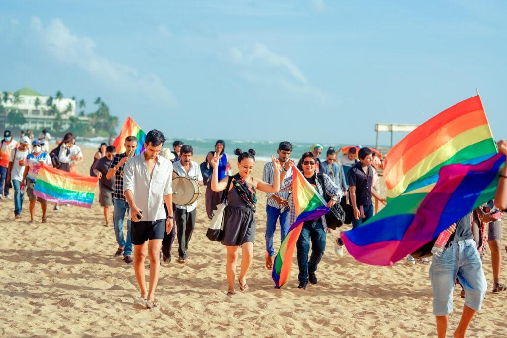 A group of people walk along a beach carrying large rainbow Pride flags
