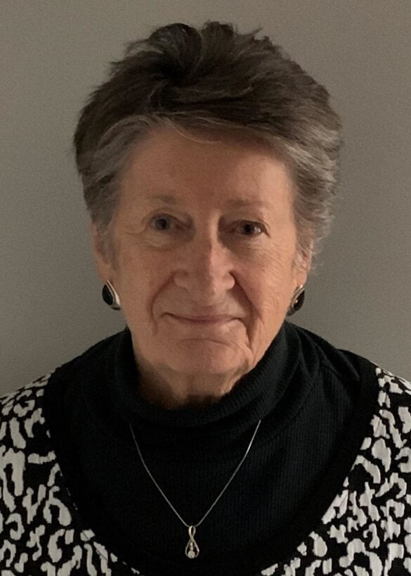 Portrait of an older woman with short grey hair, smiling, wearing a black turtleneck, a black and white patterned cardigan, and a pendant necklace.