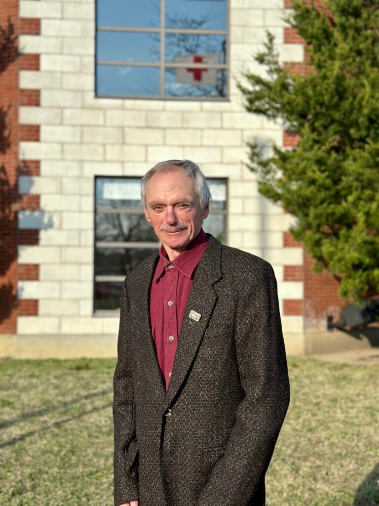 An older man with grey hair standing outdoors in front of a brick and stone building, wearing a maroon shirt and a tweed blazer with a lapel pin.