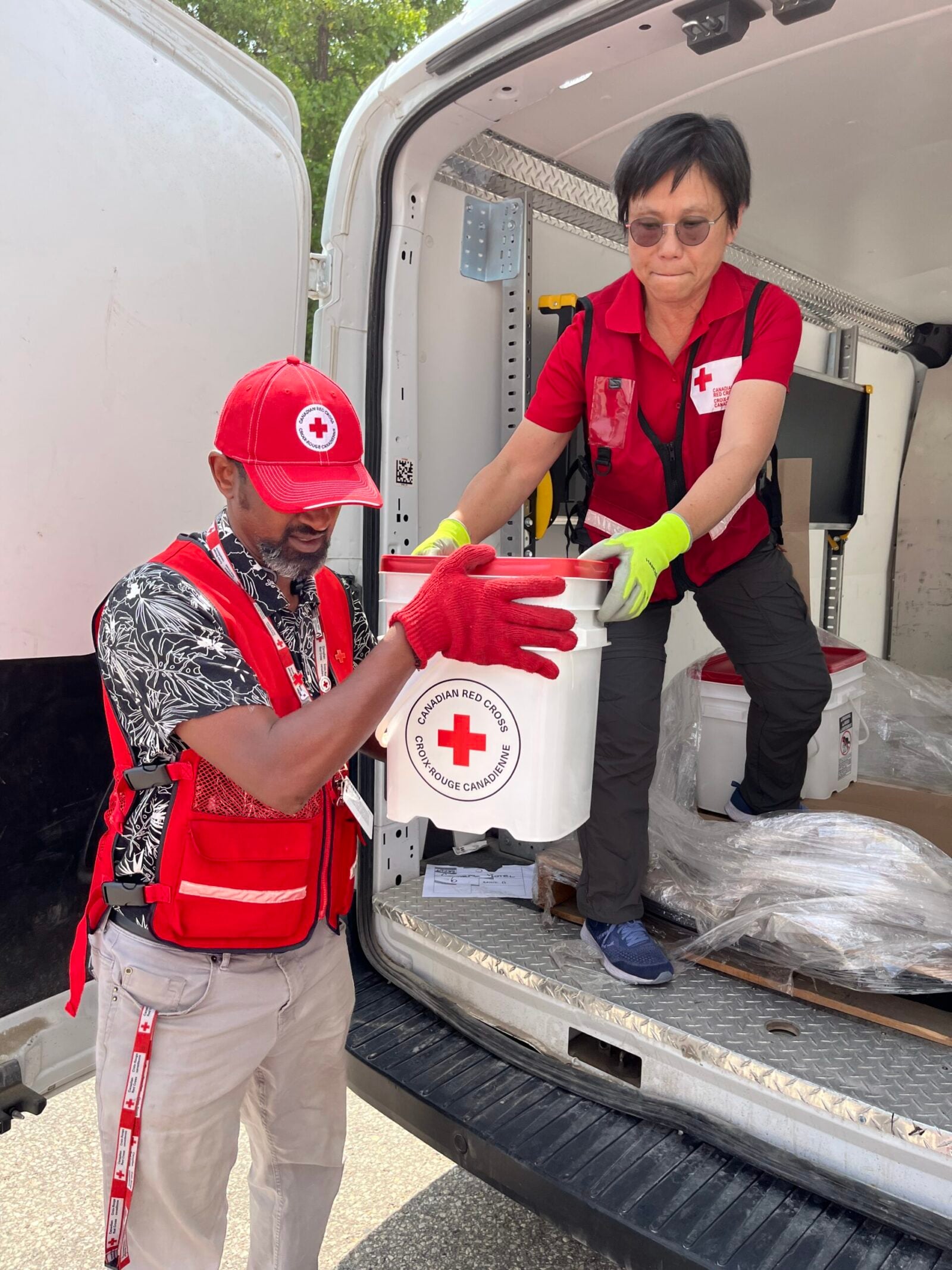 Two people wearing Red Cross logos lift a white bucket labelled 